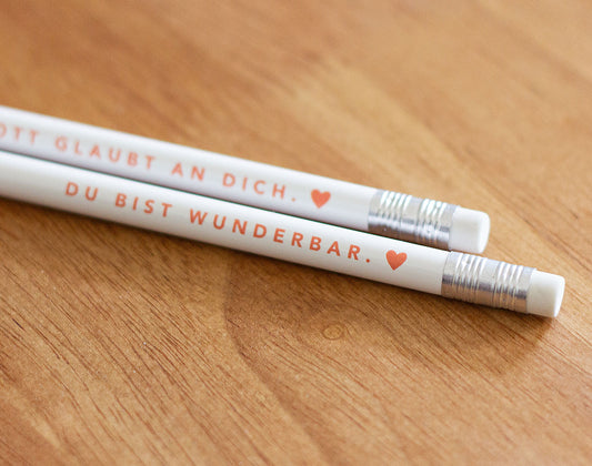 Detailaufnahme der Radiergummi-Enden: zwei weiße Bleistifte mit rotem Aufdruck „Gott glaubt an dich. ♥" und „Du bist wunderbar. ♥" – 2er-Set christliche Bleistifte aus der himmlisch-Kollektion von herzseiten.com.