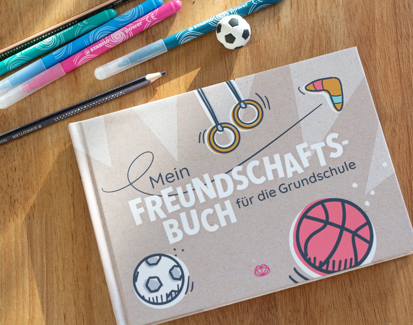 Des Buch "Mein Freundschaftsbuch für die Grundschule" liegt auf einem Tisch. Daneben eine handvoll Stifte und ein Radiergummi in Form eines Fußballs