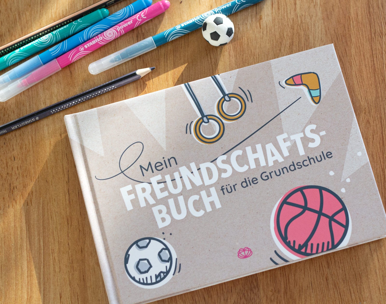 Des Buch "Mein Freundschaftsbuch für die Grundschule" liegt auf einem Tisch. Daneben eine handvoll Stifte und ein Radiergummi in Form eines Fußballs