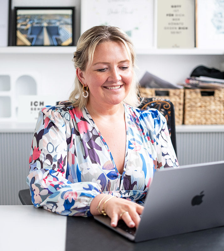 Ein Portrait von Michelle Engel, wie sie in ihrem Büro am Laptop arbeitet. Im Hintergrund sieht man unscharf verschiedene Postkarten und Poster.