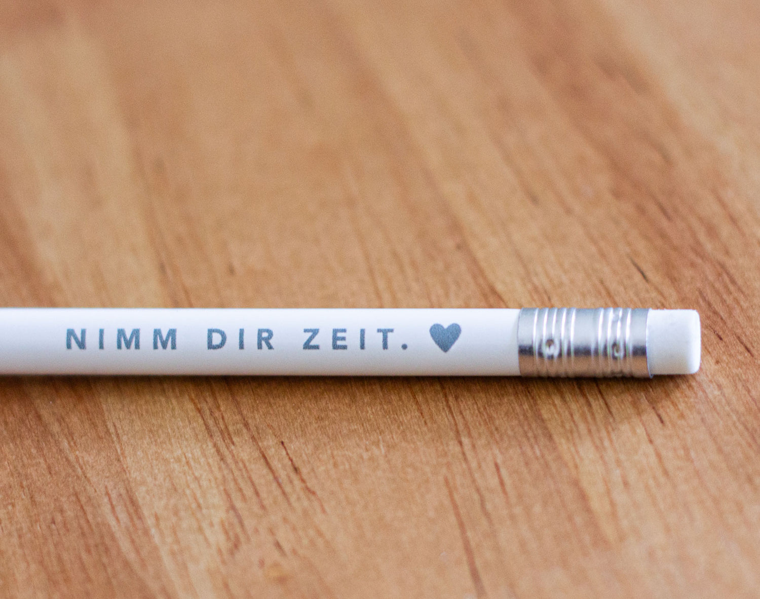 Weißer Bleistift mit grauem Aufdruck „Nimm dir Zeit. ♥" liegend auf warmem Holzuntergrund – christlicher Bleistift passend zum Buch „Nimm dir Zeit für dich und mich. Gott." von Michelle Engel, herzseiten.com.