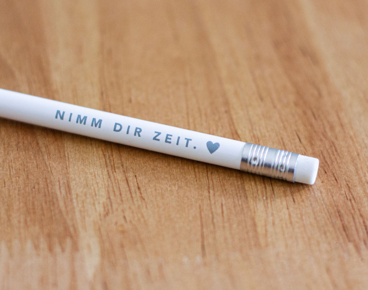 Detailaufnahme eines weißen Bleistifts mit grauem Aufdruck „Nimm dir Zeit. ♥" auf warmem Holzuntergrund – christlicher Bleistift aus der „Nimm dir Zeit"-Serie von herzseiten.com.