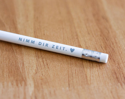 Detailaufnahme eines weißen Bleistifts mit grauem Aufdruck „Nimm dir Zeit. ♥" auf warmem Holzuntergrund – christlicher Bleistift aus der „Nimm dir Zeit"-Serie von herzseiten.com.