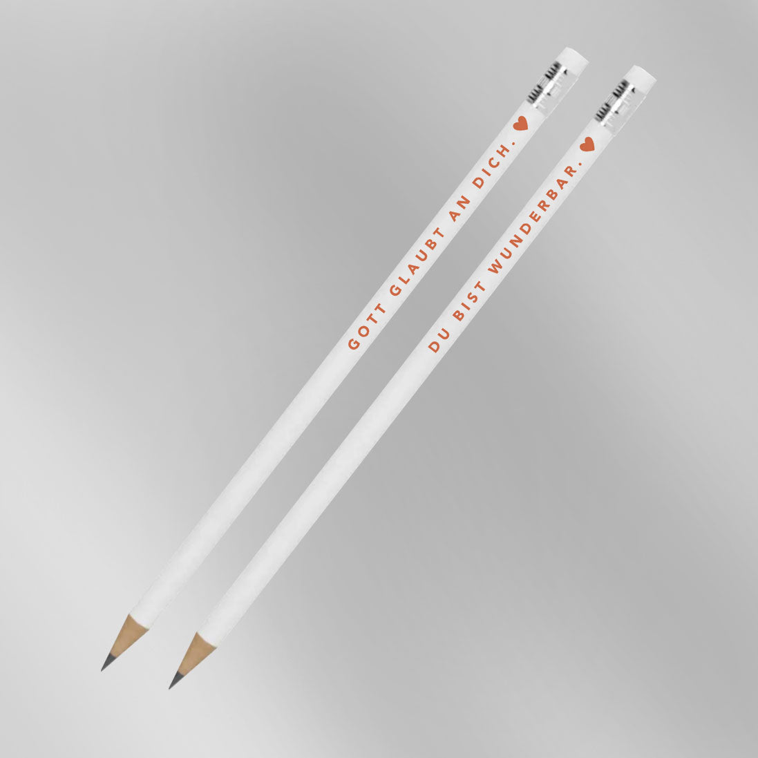 Zwei weiße Bleistifte mit Aufdruck „Gott glaubt an dich. ♥" und „Du bist wunderbar. ♥" vor grauem Hintergrund – 2er-Set christliche Bleistifte aus der himmlisch-Kollektion von herzseiten.com.