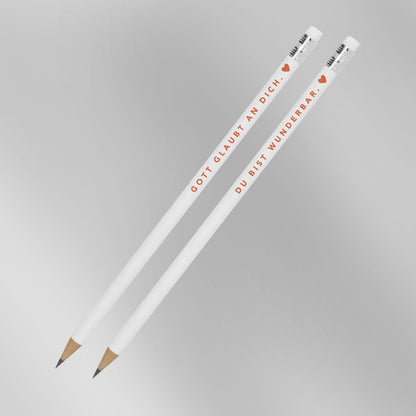Zwei weiße Bleistifte mit Aufdruck „Gott glaubt an dich. ♥" und „Du bist wunderbar. ♥" vor grauem Hintergrund – 2er-Set christliche Bleistifte aus der himmlisch-Kollektion von herzseiten.com.