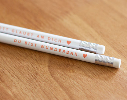 Detailaufnahme der Radiergummi-Enden: zwei weiße Bleistifte mit rotem Aufdruck „Gott glaubt an dich. ♥" und „Du bist wunderbar. ♥" – 2er-Set christliche Bleistifte aus der himmlisch-Kollektion von herzseiten.com.