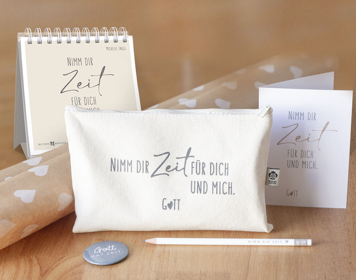 Kollektionsbild „Nimm dir Zeit für dich und mich. Gott" – alle neuen Produkte im Überblick: aufgestelltes Spiralbuch von Michelle Engel (Butzon & Bercker), naturfarbenes Baumwolltäschchen mit Organic Cotton Label, Klappkarte, weißer Bleistift „Nimm dir Zeit." und grauer Magnetbutton „Gott hat Zeit." auf Holzuntergrund mit Kraftpapier – Teil der himmlisch-Kollektion von herzseiten.com.
