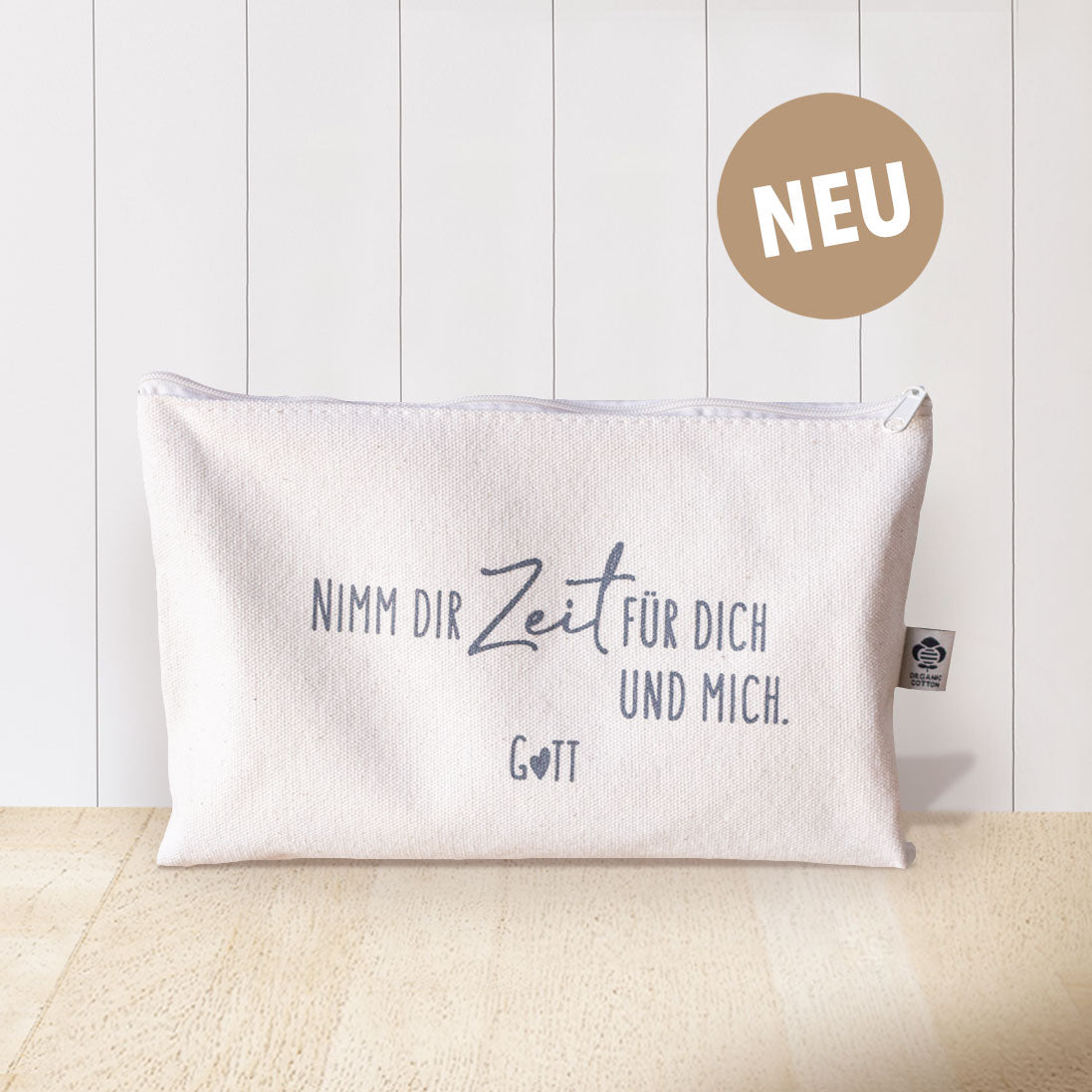 Naturfarbenes Baumwolltäschchen mit grauem Aufdruck „Nimm dir Zeit für dich und mich. Gott" und Organic Cotton Label vor weißem Hintergrund mit goldenem NEU-Badge – christliches Täschchen aus der himmlisch-Kollektion von herzseiten.com.