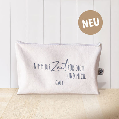 Naturfarbenes Baumwolltäschchen mit grauem Aufdruck „Nimm dir Zeit für dich und mich. Gott" und Organic Cotton Label vor weißem Hintergrund mit goldenem NEU-Badge – christliches Täschchen aus der himmlisch-Kollektion von herzseiten.com.