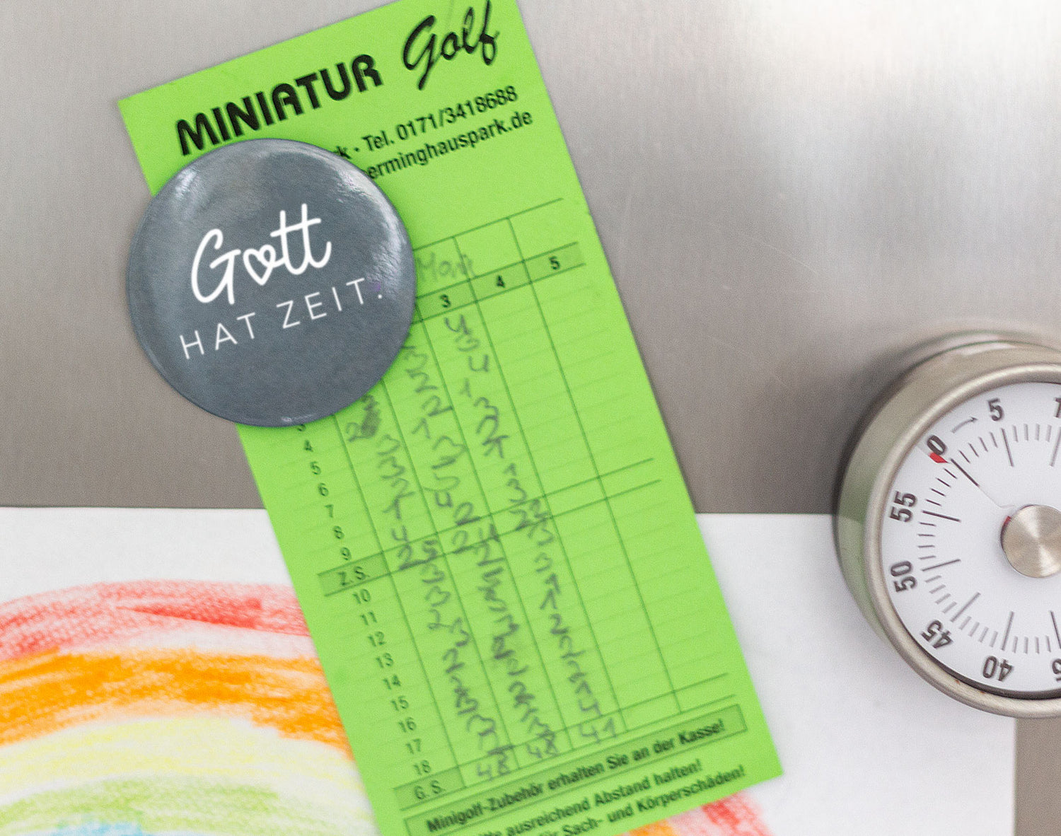 Magnetbutton „Gott hat Zeit." in Dunkelgrau mit weißer Schrift, befestigt an einem Kühlschrank neben einem Küchentimer und einer Kinderzeichnung mit Regenbogen. Im Hintergrund ein Minigolf-Scorezettel.