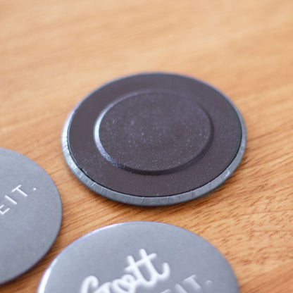 Nahaufnahme der Rückseite eines runden Magnetbuttons mit schwarzem Magneteinsatz auf Holzuntergrund. Im Vordergrund und an der Seite sind weitere Buttons mit der Aufschrift „Gott hat Zeit." erkennbar.
