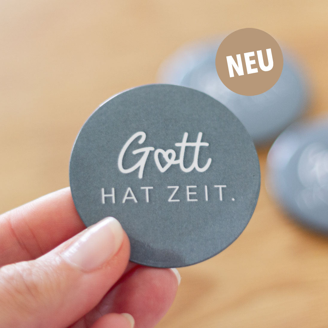 Eine Hand hält einen runden Magnetbutton in Dunkelgrau mit der Aufschrift „Gott hat Zeit." in weißer Schrift. Im Hintergrund zwei weitere Buttons auf hellem Holz. Oben rechts ein beiger „NEU"-Badge.