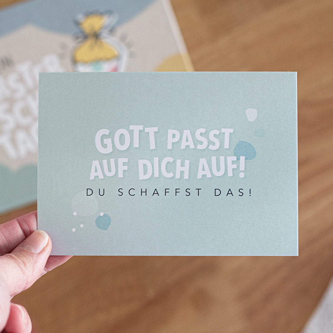 Eine Hand hält die Postkarte "Gott passt auf dich auf! Du schaffst das!"