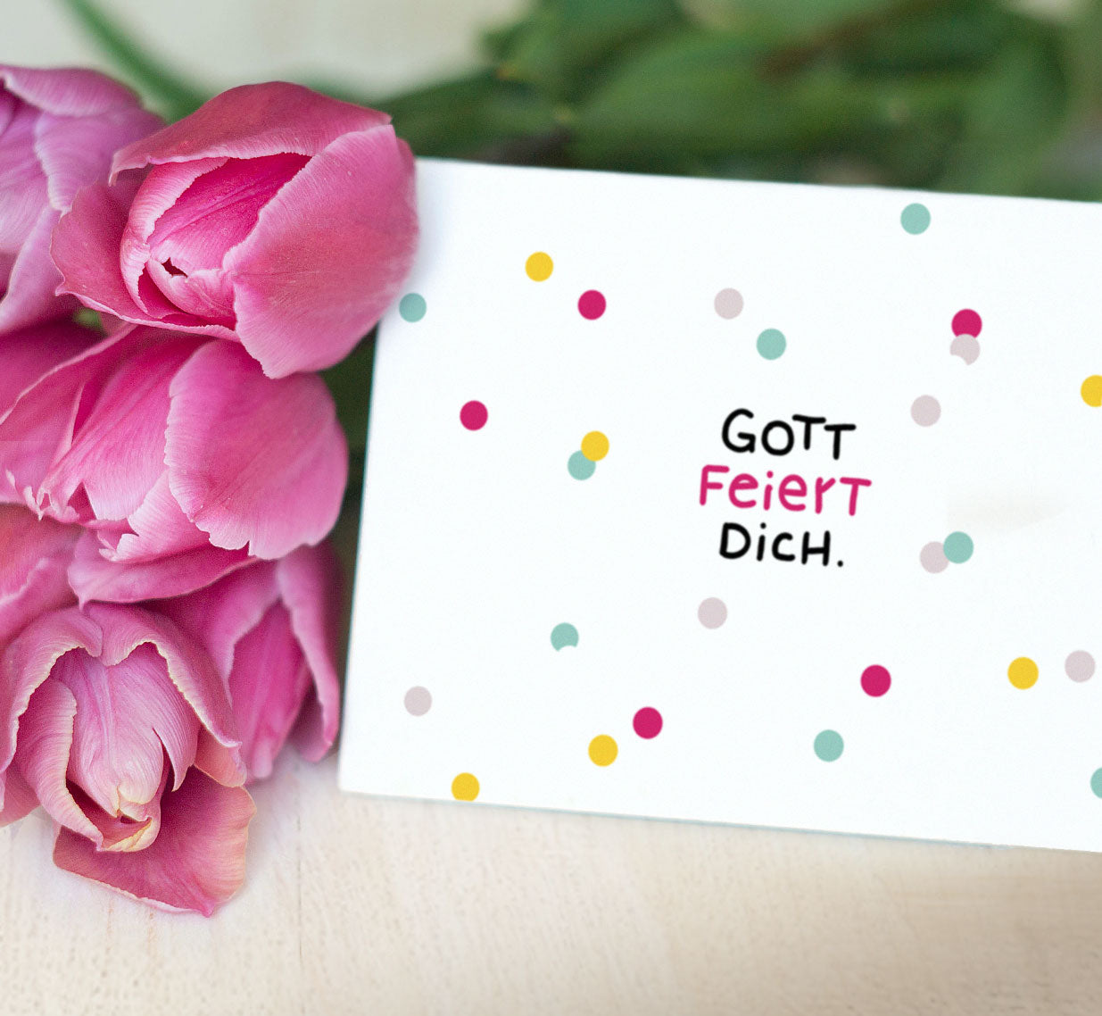 Christliche Postkarte „Gott feiert dich." mit bunten Konfetti-Punkten, neben pinken Tulpen auf hellem Holzuntergrund – aus dem Postkartenset „Gott hat dich wow gemacht." 