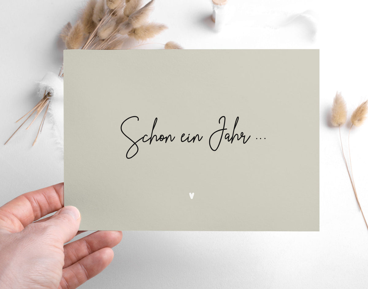 Postkarte „Schon ein Jahr …" in warmem Greige mit schwarzer Kalligrafie und weißem Herzchen, in einer Hand gehalten vor Trockenblumen-Arrangement – aus der himmlisch-Kollektion von herzseiten.com.