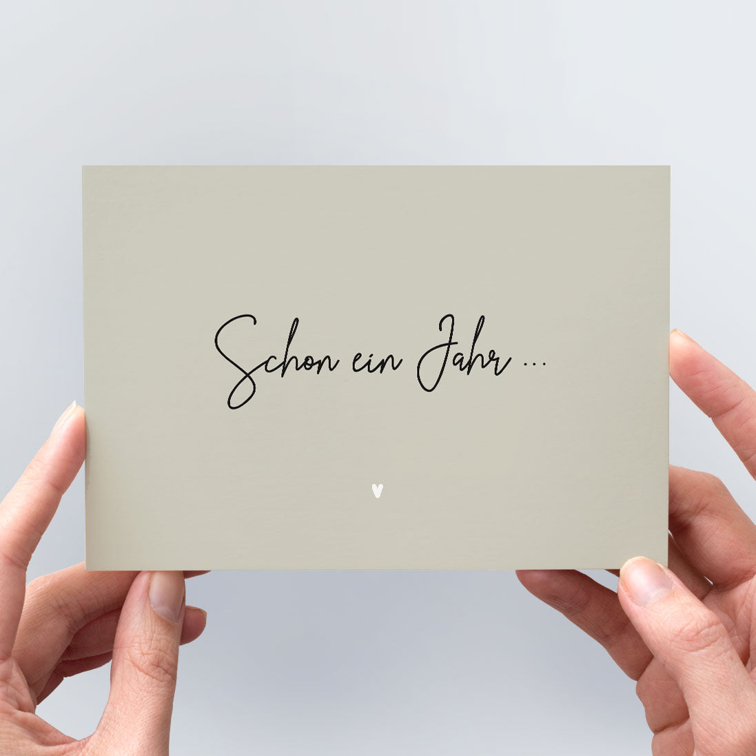 Postkarte „Schon ein Jahr …" in warmem Greige mit schwarzer Kalligrafie und weißem Herzchen, in zwei Händen gehalten vor hellem Hintergrund – minimalistische Grußkarte aus der himmlisch-Kollektion von herzseiten.com.