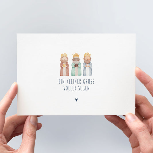 Postkarte „Ein kleiner Gruß voller Segen" mit zarter Aquarell-Illustration der Heiligen Drei Könige in Erdtönen, marineblauer Typografie und kleinem Herzchen, in zwei Händen gehalten vor hellem Hintergrund – christliche Weihnachtspostkarte aus der himmlisch-Kollektion von herzseiten.com.