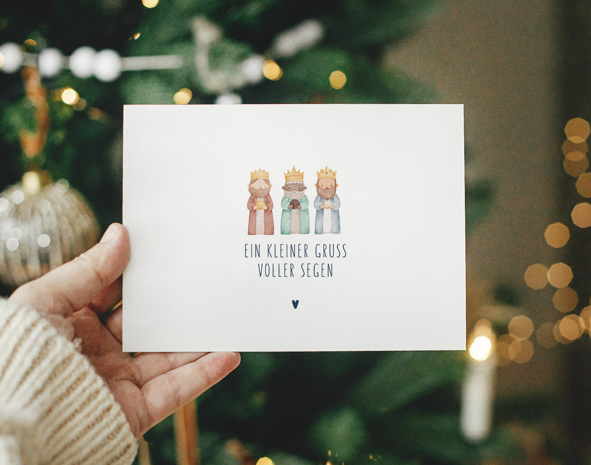 Postkarte „Ein kleiner Gruß voller Segen" mit Aquarell-Illustration der Heiligen Drei Könige und marineblauer Typografie, in einer Hand gehalten vor beleuchtetem Weihnachtsbaum – christliche Weihnachtspostkarte aus der himmlisch-Kollektion von herzseiten.com.