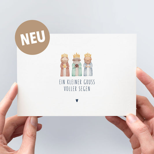 Postkarte „Ein kleiner Gruß voller Segen" mit zarter Aquarell-Illustration der Heiligen Drei Könige in Erdtönen, marineblauer Typografie und kleinem Herzchen, in zwei Händen gehalten vor hellem Hintergrund – christliche Weihnachtspostkarte aus der himmlisch-Kollektion mit neu-Badge