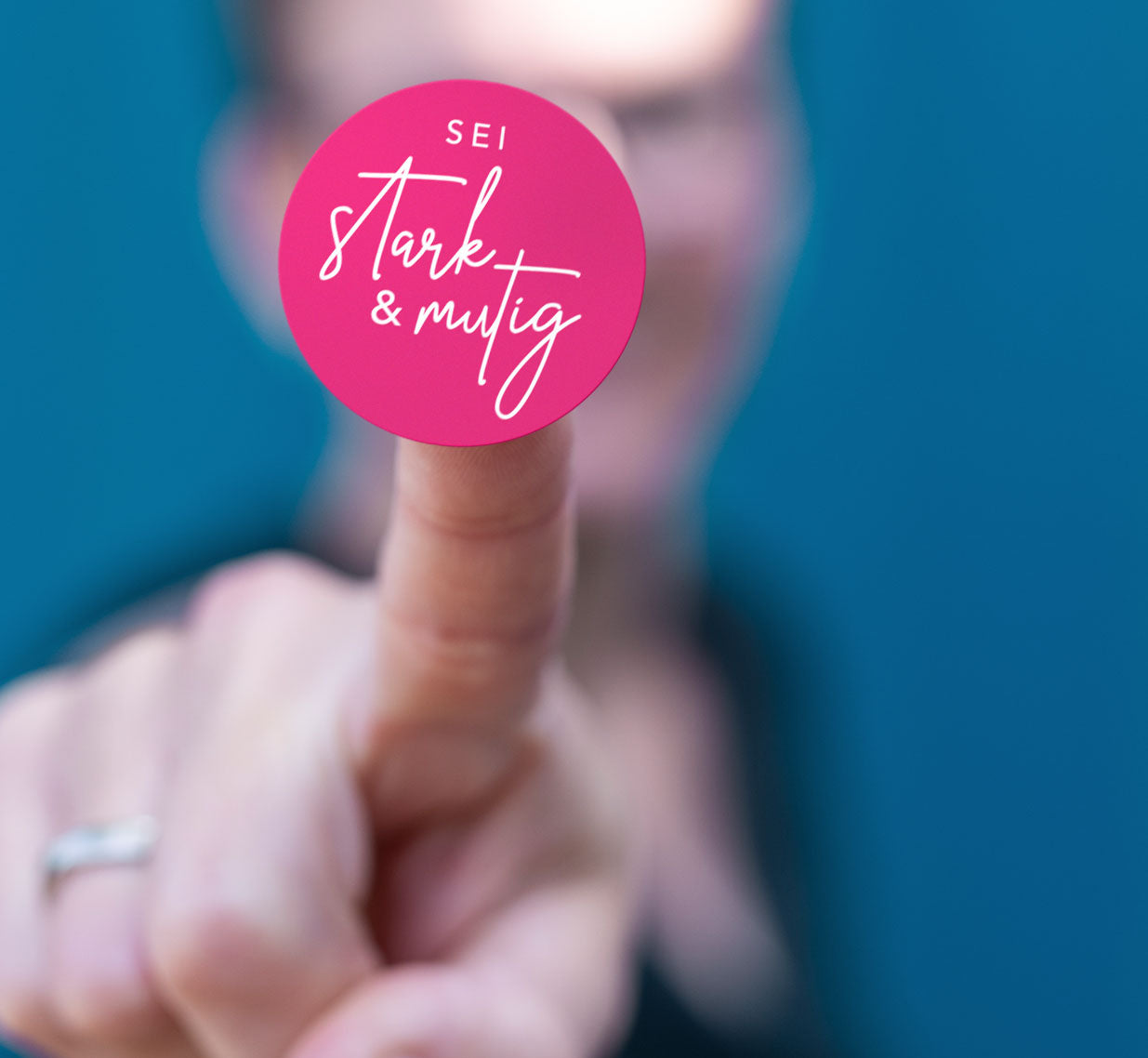 Pinker runder Sticker mit weißer Handschrift „Sei stark & mutig" auf einem Finger. Detailmotiv aus dem christlichen Stickerbogen „Gott hat dich wow gemacht" der himmlisch-Kollektion von herzseiten.com.