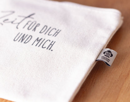 Detailaufnahme des naturfarben Baumwolltäschchens mit grauem Aufdruck „Zeit für dich und mich." und eingenähtem Organic Cotton Label – Täschchen aus der „Nimm dir Zeit"-Serie von herzseiten.com.